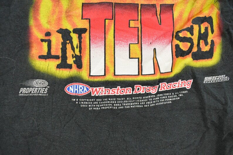Op de afbeelding: Zwart T-shirt met een opvallende afbeelding van "iNTENSE" in rode en witte letters tegen een gele en oranje vlammenachtergrond. Voorzien van "NHRA Winston Drag Racing" branding.