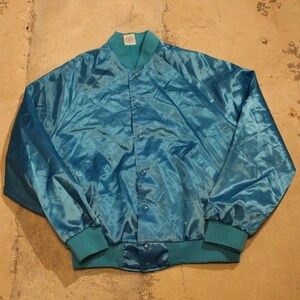May include: A teal satin bomber jacket with a button-up front, teal ribbed collar, cuffs, and hem. The jacket is a solid color and appears to be vintage. The jacket is laying flat on a concrete surface.