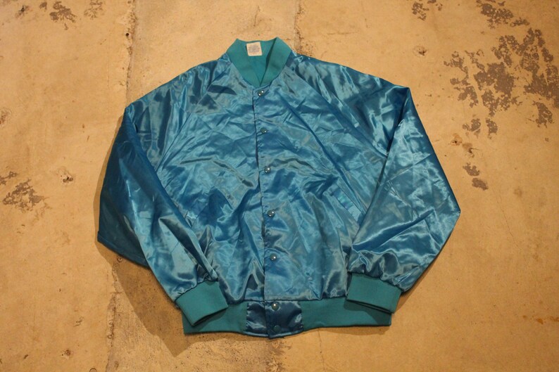 May include: A teal satin bomber jacket with a button-up front, teal ribbed collar, cuffs, and hem. The jacket is a solid color and appears to be vintage. The jacket is laying flat on a concrete surface.