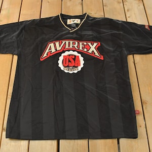 Op de afbeelding: Zwart shirt met korte mouwen en verticale strepen. Het shirt heeft het woord "AVIREX" in rood en wit over de borst, boven een wit en rood "USA" embleem. De kraag is goudkleurig en de mouwen hebben rode en witte randen.