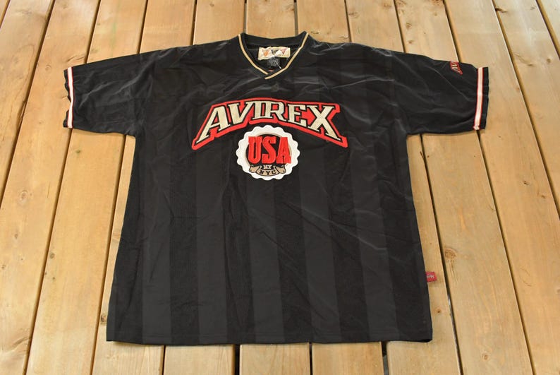 Op de afbeelding: Zwart shirt met korte mouwen en verticale strepen. Het shirt heeft het woord "AVIREX" in rood en wit over de borst, boven een wit en rood "USA" embleem. De kraag is goudkleurig en de mouwen hebben rode en witte randen.