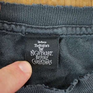 May include: Close-up of a dark gray t-shirt collar with a black tag. The tag features the text "Disney Tim Burton's The Nightmare Before Christmas" in white lettering, indicating a themed apparel item.