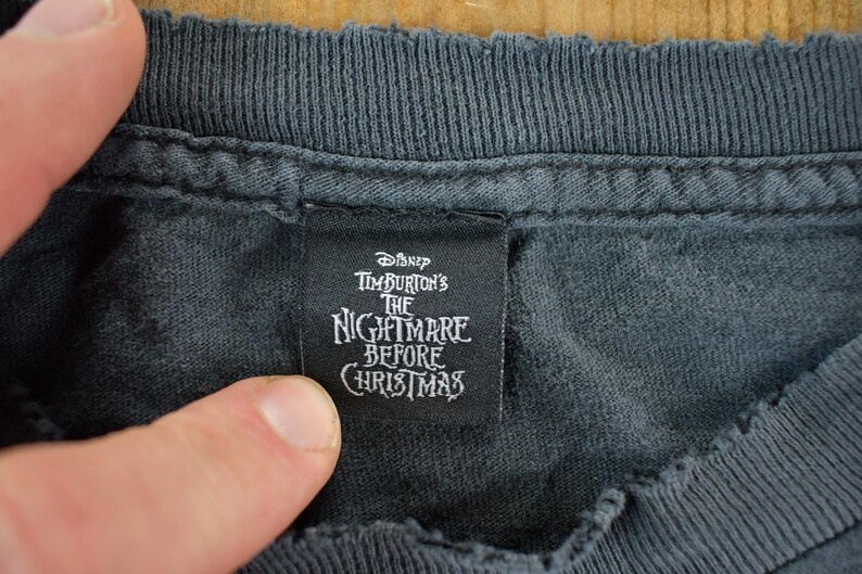 May include: Close-up of a dark gray t-shirt collar with a black tag. The tag features the text "Disney Tim Burton's The Nightmare Before Christmas" in white lettering, indicating a themed apparel item.