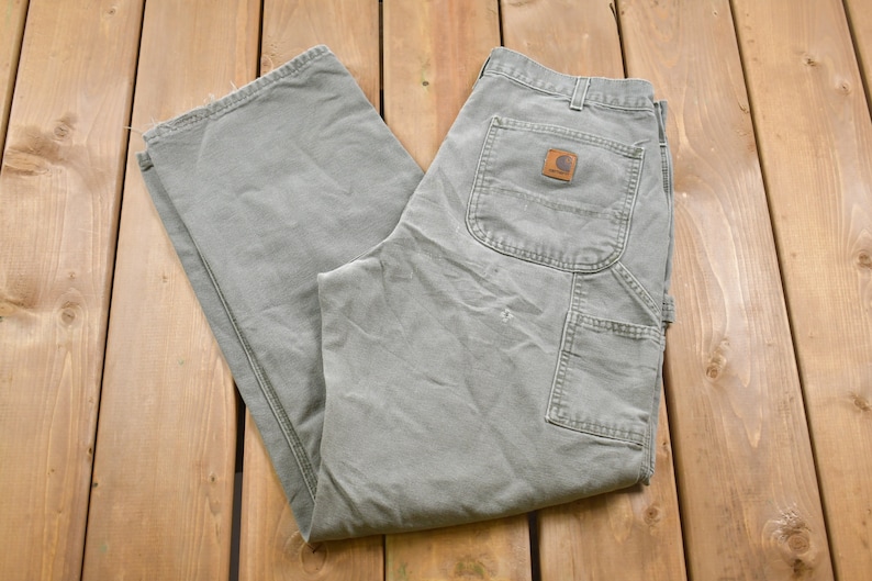 May include: A pair of grey work trousers with a brown Carhartt logo patch on the back pocket.