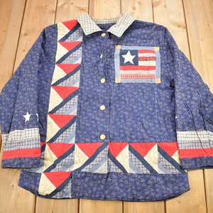 May include: A blue long-sleeved button-up shirt with a patchwork design featuring red, white and blue triangles. The shirt has a small American flag patch on the left chest.