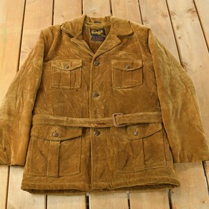 May include: A vintage brown corduroy jacket with a belted waist and multiple pockets. The jacket has a collared neckline and button closures. The corduroy fabric has a soft, textured appearance. The jacket is laid out on a wooden surface.