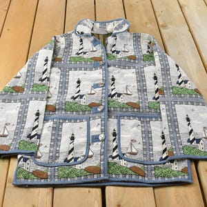 May include: A long-sleeve button-up jacket featuring a repeating tapestry-style pattern of black and white striped lighthouses, sailboats, and green grassy patches on a light gray background, complete with two large front patch pockets.