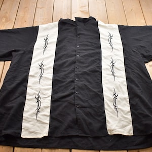 May include: A black and white short-sleeved button-up shirt with a vertical striped pattern. The shirt has black tribal-style designs on the white stripes.