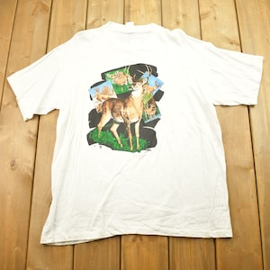 Pode incluir: Camiseta branca com uma estampa gr&aacute;fica de um veado em um cen&aacute;rio de floresta. O veado est&aacute; cercado por folhagem verde e outros veados no fundo.