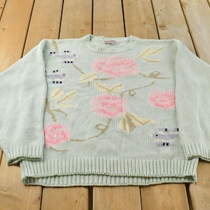 May include: A light mint green long-sleeve sweater featuring a knit pattern of pink roses, tan leaves, and small blue and white bird motifs. The garment has a ribbed crew neckline, ribbed cuffs, and a ribbed hemline with a standard pullover design.
