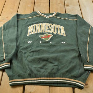 May include: A dark green long-sleeve sweatshirt featuring the text MINNESOTA in gold lettering above the Minnesota Wild logo and the word WILD in white. The garment includes ribbed cuffs and a hem with gold, white, and green striped accents.