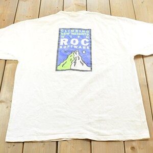 May include: White t-shirt with a graphic on the back featuring a mountain scene and the text "CLIMBING NEW HEIGHTS WITH ROC SOFTWARE". The graphic includes green, blue, and white colours. The shirt is laid flat on a wooden surface.