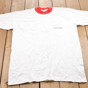 May include: White t-shirt with red trim around the collar and sleeves. The shirt has a pocket on the left side with the word "Cambridge" embroidered in red.