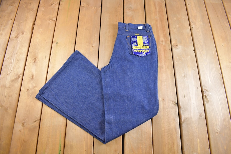 Op de afbeelding: Een paar blauwe denim jeans met een wijde pijp, plat gelegd op een houten oppervlak. De jeans heeft een geel label met de tekst "Wrangler" in blauw.