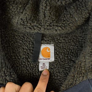 Puede incluir: Primer plano de la etiqueta de una chaqueta Carhartt. La etiqueta es blanca con el logotipo de Carhartt, una 'C' naranja dentro de un cuadrado blanco. La etiqueta dice 'XL TALL Made in Mexico'. La chaqueta tiene un interior gris oscuro y difuso.
