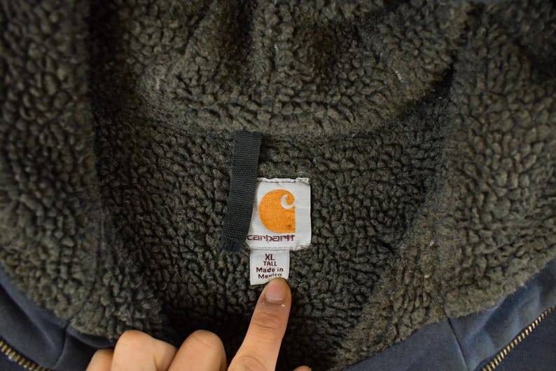 Puede incluir: Primer plano de la etiqueta de una chaqueta Carhartt. La etiqueta es blanca con el logotipo de Carhartt, una 'C' naranja dentro de un cuadrado blanco. La etiqueta dice 'XL TALL Made in Mexico'. La chaqueta tiene un interior gris oscuro y difuso.