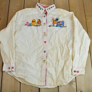 May include: A white long-sleeved button-down shirt with embroidered Winnie the Pooh characters and colourful buttons. The collar is trimmed with red and white fabric.