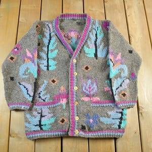 Könnte beinhalten: Graue Strickjacke mit Knöpfen, V-Ausschnitt und langen Ärmeln. Der Pullover hat ein farbenfrohes Blumen- und Blättermuster in Blau-, Lila- und Rosatönen. Die Einfassung und Knopfleiste sind rosa.