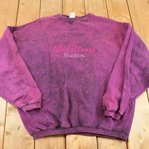 May include: A long-sleeve crewneck sweatshirt in a deep magenta acid-wash finish. The center chest features the embroidered text The Walt Disney Studios in a matching magenta thread. The garment has ribbed cuffs and a ribbed hemline.