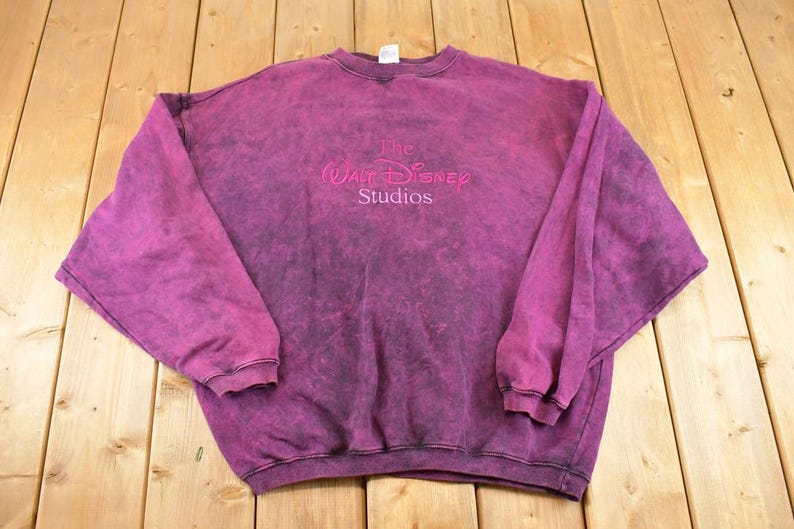 May include: A long-sleeve crewneck sweatshirt in a deep magenta acid-wash finish. The center chest features the embroidered text The Walt Disney Studios in a matching magenta thread. The garment has ribbed cuffs and a ribbed hemline.