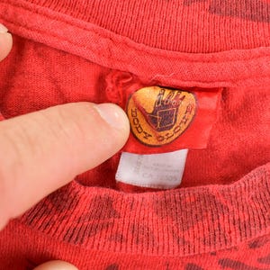 May include: Close-up of a red t-shirt with a dark pattern. The shirt's tag features the "Body Glove" logo in gold and black. The tag is attached to the collar of the shirt. The shirt is made of a soft material.
