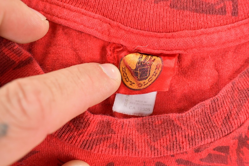 May include: Close-up of a red t-shirt with a dark pattern. The shirt's tag features the "Body Glove" logo in gold and black. The tag is attached to the collar of the shirt. The shirt is made of a soft material.