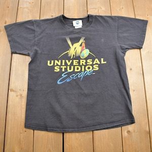 Może przedstawiać: Ciemnoszara koszulka z logo Universal Studios i napisem "UNIVERSAL STUDIOS Escape" w kolorze żółtym i niebieskim. Koszulka leży płasko na drewnianej powierzchni.