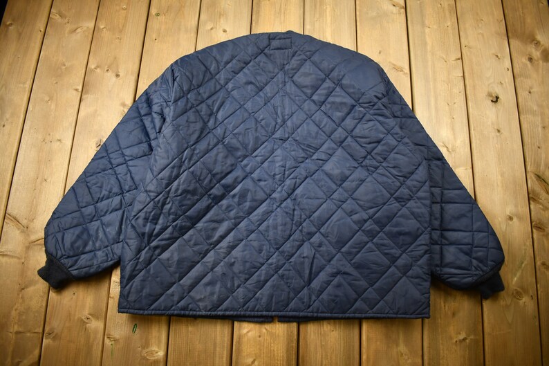 May include: A navy blue quilted jacket with a diamond pattern. The jacket has a zipper closure and a stand-up collar.