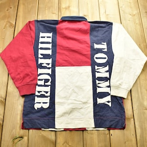 Pode incluir: Uma camisa de rugby de manga comprida com um design de blocos de cores em vermelho, branco e azul. A camisa tem as palavras "Tommy" e "Hilfiger" impressas nas mangas em branco.
