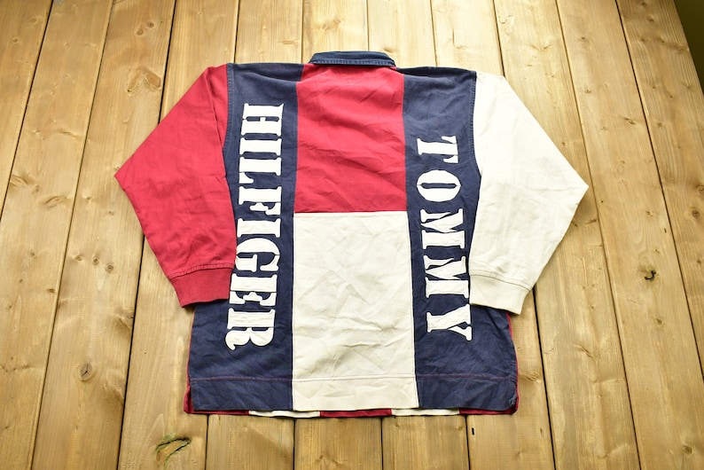 Pode incluir: Uma camisa de rugby de manga comprida com um design de blocos de cores em vermelho, branco e azul. A camisa tem as palavras "Tommy" e "Hilfiger" impressas nas mangas em branco.