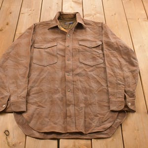 May include: A brown button-down shirt with two chest pockets. The long-sleeved shirt has a collar and buttoned cuffs. The fabric has a textured appearance. The shirt is laid flat on a wooden surface.