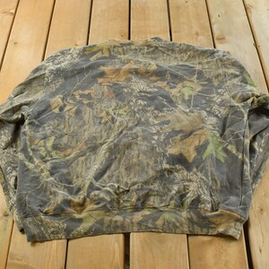May include: A camouflage-patterned sweatshirt in shades of green, brown, and grey, laid flat on a wooden surface. The design features a mix of leaf and branch patterns, typical of hunting apparel.