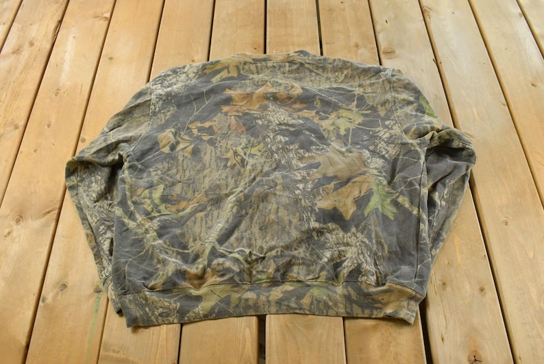 May include: A camouflage-patterned sweatshirt in shades of green, brown, and grey, laid flat on a wooden surface. The design features a mix of leaf and branch patterns, typical of hunting apparel.