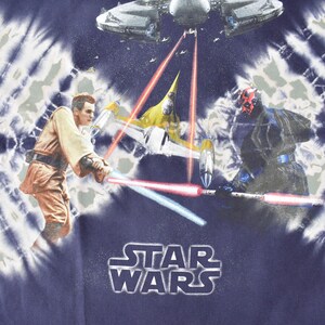 May include: A blue tie-dye t-shirt with a Star Wars theme. The shirt features a scene from the movie with characters from the Star Wars universe, including a Jedi, a Sith Lord, and a spaceship. The text "STAR WARS" is printed on the front of the shirt.