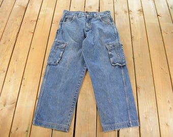 May include: Light blue denim cargo pants with multiple pockets, including two large side pockets. The pants have a classic five-pocket design and a button and zipper closure. The denim has a faded, worn look.