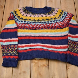 May include: A colorful striped sweater with a geometric pattern around the neck. The sweater is made of a soft, knitted fabric and features a variety of colors, including blue, red, yellow, pink, and white.