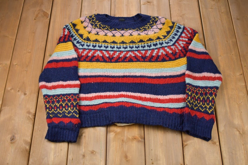 May include: A colorful striped sweater with a geometric pattern around the neck. The sweater is made of a soft, knitted fabric and features a variety of colors, including blue, red, yellow, pink, and white.