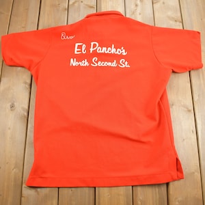 May include: Red short-sleeved polo shirt with white embroidered text on the back that reads "El Pancho's North Second St."
