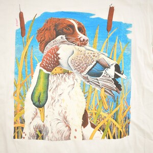 May include: A white t-shirt with a colorful illustration of a brown and white dog holding a duck in its mouth. The dog is standing in tall grass with blue sky in the background.