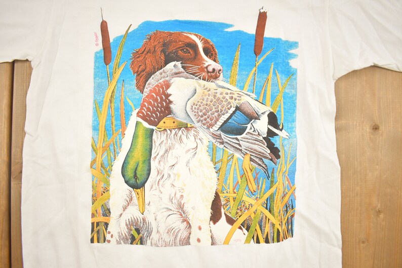 May include: A white t-shirt with a colorful illustration of a brown and white dog holding a duck in its mouth. The dog is standing in tall grass with blue sky in the background.