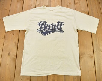Könnte beinhalten: Ein weißes T-Shirt mit einem blauen und weißen "Banff"-Aufdruck auf der Vorderseite.