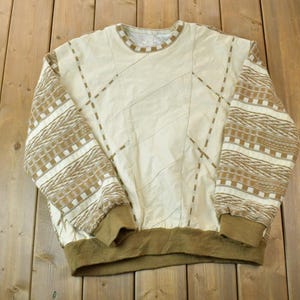 May include: A cream-colored sweater with brown and white geometric patterns on the sleeves. The sweater has a crew neck and a ribbed hem.