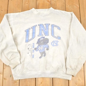 K&ouml;nnte beinhalten: Hellgraues Sweatshirt mit den Buchstaben "UNC" in Hellblau auf der Brust. Das Sweatshirt zeigt ein Cartoon-Tar Heel-Maskottchen und andere Grafiken. Das Sweatshirt ist aus weichem Material und hat lange &Auml;rmel.
