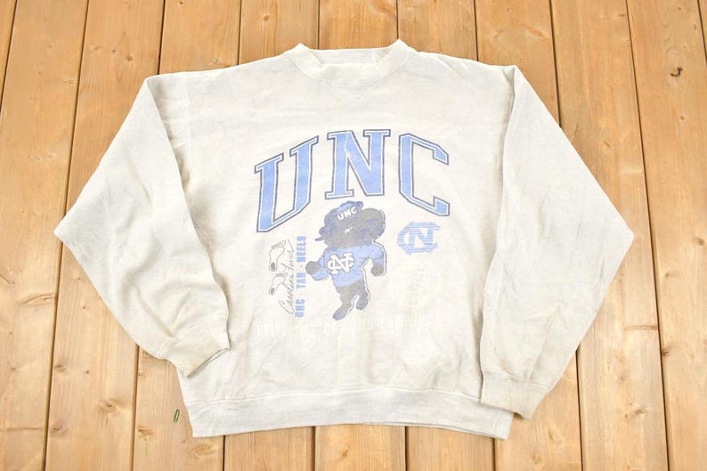 K&ouml;nnte beinhalten: Hellgraues Sweatshirt mit den Buchstaben "UNC" in Hellblau auf der Brust. Das Sweatshirt zeigt ein Cartoon-Tar Heel-Maskottchen und andere Grafiken. Das Sweatshirt ist aus weichem Material und hat lange &Auml;rmel.