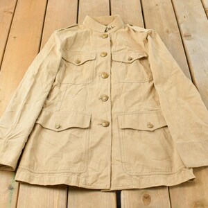 May include: Tan jacket with a mandarin collar, four front pockets, and button closures. The jacket has long sleeves and epaulets on the shoulders. The jacket is laid out on a wooden surface.