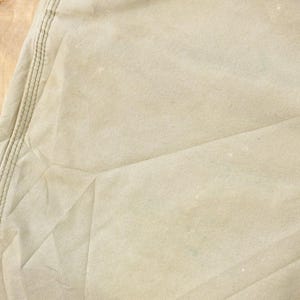 May include: Close-up of a section of tan fabric, possibly canvas or a similar material. Visible are seams with stitching, and the fabric has a slightly worn appearance with some light-colored spots.