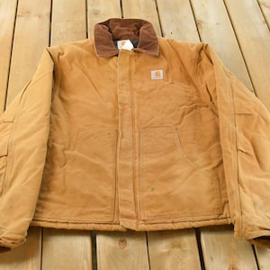 Puede incluir: Chaqueta de trabajo de color marr&oacute;n claro con cuello de pana marr&oacute;n. La chaqueta tiene dos bolsillos delanteros y un peque&ntilde;o logotipo cuadrado en el pecho. La chaqueta est&aacute; sobre una superficie de madera.