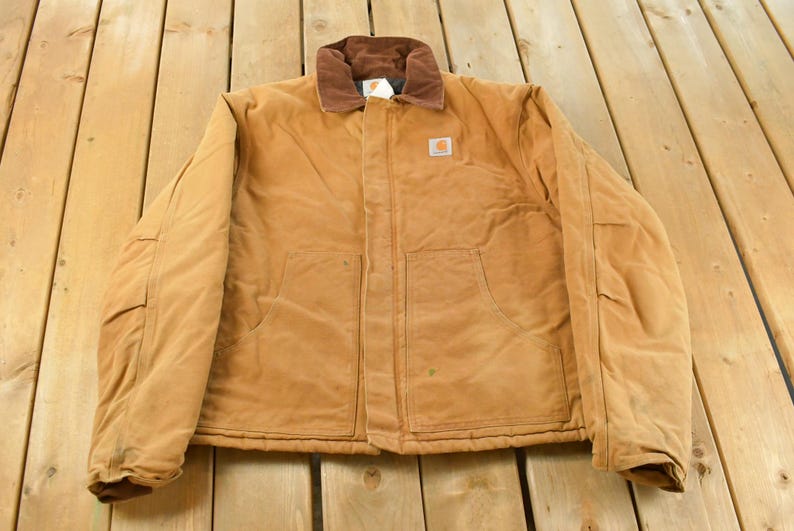 Puede incluir: Chaqueta de trabajo de color marr&oacute;n claro con cuello de pana marr&oacute;n. La chaqueta tiene dos bolsillos delanteros y un peque&ntilde;o logotipo cuadrado en el pecho. La chaqueta est&aacute; sobre una superficie de madera.