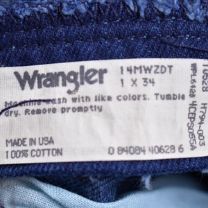May include: A white Wrangler jeans care label with the text "Wrangler", "14MWZDT", "1 X 34", "WPL6428 4CEPS055A", "10528 H794-003", "Machine wash with like colors. Tumble dry. Remove promptly", "MADE IN USA", "100% COTTON", and "0 84084 40628 6".