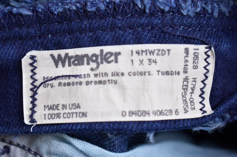 May include: A white Wrangler jeans care label with the text "Wrangler", "14MWZDT", "1 X 34", "WPL6428 4CEPS055A", "10528 H794-003", "Machine wash with like colors. Tumble dry. Remove promptly", "MADE IN USA", "100% COTTON", and "0 84084 40628 6".